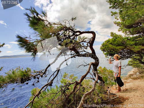 Image of Young active feamle tourist taking a break, drinking water, wearing small backpack while walking on coastal path among pine trees looking for remote cove to swim alone in peace on seaside in Croatia