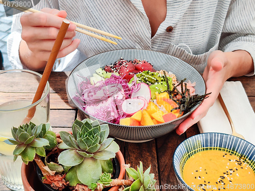 Image of Woman eating tasty colorful healthy natural organic vegetarian Hawaiian poke bowl using asian chopsticks on rustic wooden table. Healthy natural organic eating concept