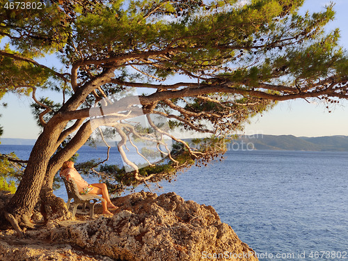 Image of Pensive woman on vacations, sitting and relaxing under large pine tree on bench by dip blue sea enjoying beautiful sunset light in Brela, Makarska region, Dalmatia, Croatia