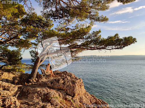 Image of Pensive woman on vacations, sitting and relaxing under large pine tree on bench by dip blue sea enjoying beautiful sunset light in Brela, Makarska region, Dalmatia, Croatia