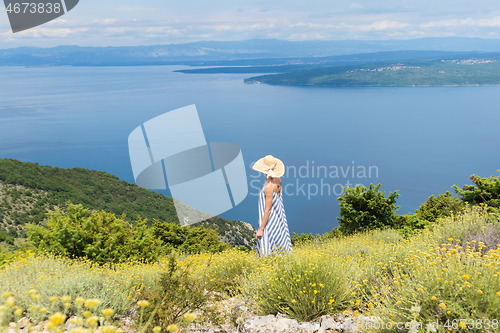 Image of Rear view of young woman wearing striped summer dress and straw hat standing in super bloom of wildflowers, relaxing while enjoing beautiful view of Adriatic sea nature, Croatia