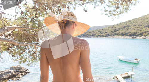 Image of Rear view of topless beautiful woman wearing nothing but straw sun hat realaxing on wild coast of Adriatic sea on a beach in shade of pine tree.