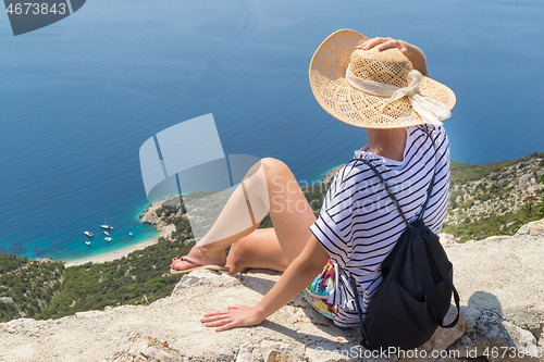 Image of Active sporty woman on summer vacations sitting on old stone wall at Lubenice village, wearing straw hat and beach backpack enjoying beautiful coastal view of Cres island, Croatia