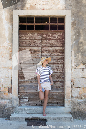 Image of Beautiful young female tourist woman standing in front of vinatage wooden door and textured stone wall at old Mediterranean town, smiling, holding, smart phone to network on vacationes