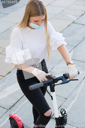 Image of Trendy fashinable girl wearing corona virus protective face mask and rubber gloves while using rental electric scooters in city environment. Eco-friendly city transport in Ljubljana, Slovenia