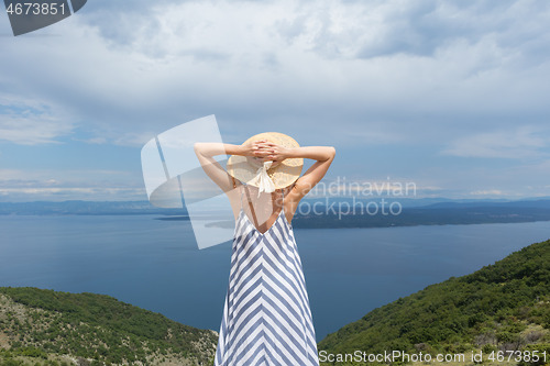 Image of Rear view of young woman wearing striped summer dress and straw hat standing in super bloom of wildflowers, relaxing while enjoing beautiful view of Adriatic sea nature, Croatia