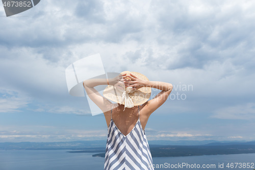 Image of Rear view of young woman wearing striped summer dress and straw hat standing in super bloom of wildflowers, relaxing while enjoing beautiful view of Adriatic sea nature, Croatia