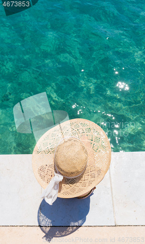 Image of Woman wearing big summer sun hat relaxing on pier by clear turquoise sea.