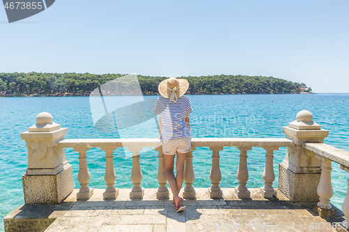 Image of Rear view of woman wearing straw summer hat ,leaning against elegant old stone fence of coastal villa, relaxing while looking at blue Adriatic sea, on Losinj island Croatia.