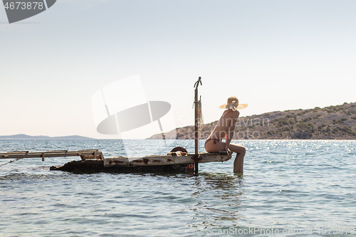 Image of View of unrecognizable woman wearing big summer sun hat tanning topless and relaxing on old wooden pier in remote calm cove of Adriatic sea, Croatia