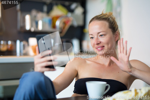 Image of Young smiling cheerful pleased woman indoors at home kitchen using social media on mobile phone for chatting and staying connected with her loved ones. Stay at home, social distancing lifestyle.