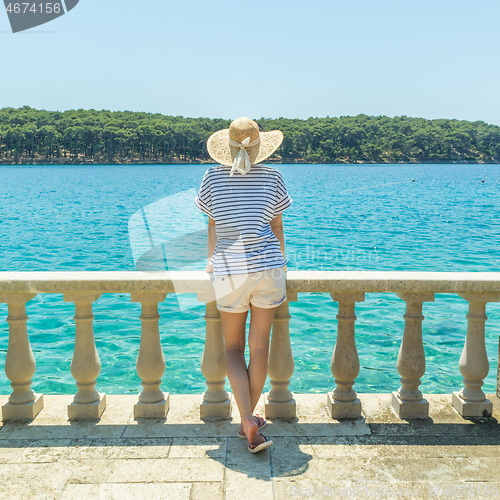 Image of Rear view of woman wearing straw summer hat ,leaning against elegant old stone fence of coastal villa, relaxing while looking at blue Adriatic sea, on Losinj island Croatia.