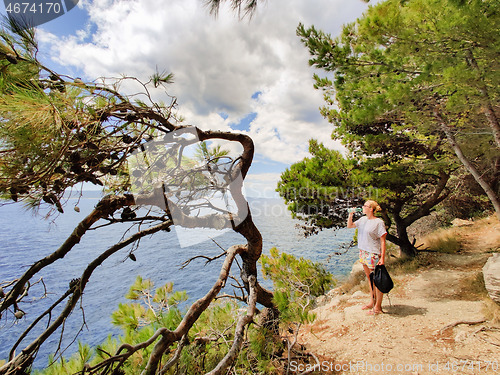 Image of Young active feamle tourist taking a break, drinking water, wearing small backpack while walking on coastal path among pine trees looking for remote cove to swim alone in peace on seaside in Croatia