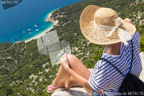 Image of Active sporty woman on summer vacations sitting on old stone wall at Lubenice village, wearing straw hat and beach backpack enjoying beautiful coastal view of Cres island, Croatia