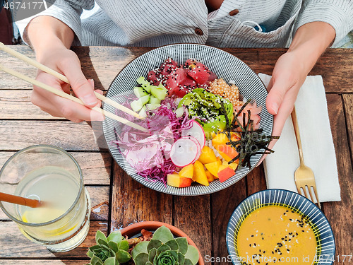 Image of Woman eating tasty colorful healthy natural organic vegetarian Hawaiian poke bowl using asian chopsticks on rustic wooden table. Healthy natural organic eating concept