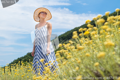 Image of Young woman wearing striped summer dress and straw hat standing in super bloom of wildflowers, relaxing while enjoing beautiful nature of of Adriatic sea coastal nature of Croatia.