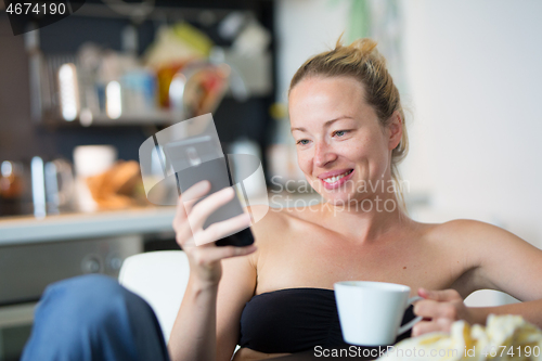 Image of Young smiling cheerful pleased woman indoors at home kitchen using social media on mobile phone for chatting and staying connected with her loved ones. Stay at home, social distancing lifestyle.