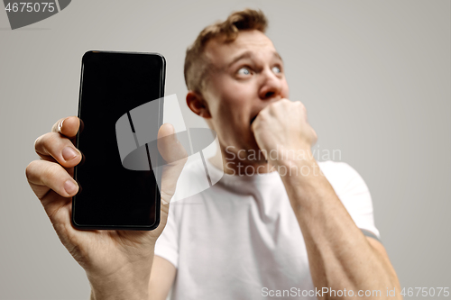 Image of Young handsome man showing smartphone screen isolated on gray background in shock with a surprise face