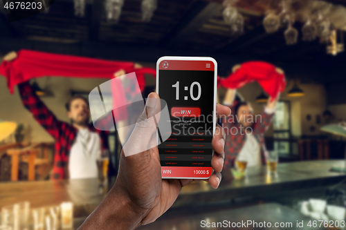 Image of Device screen with mobile app for betting and score, cheering friends, fans on background