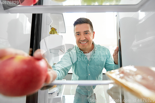 Image of man taking apple from fridge at home kitchen