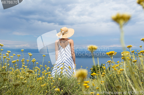 Image of Rear view of young woman wearing striped summer dress and straw hat standing in super bloom of wildflowers, relaxing while enjoing beautiful view of Adriatic sea nature, Croatia