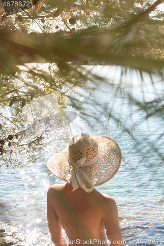 Image of Rear view of topless beautiful woman wearing nothing but straw sun hat realaxing on wild coast of Adriatic sea on a beach in shade of pine tree.