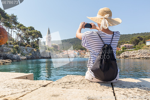 Image of Woman traveler wearing straw summer hat and backpack, sittingat edge of stone pier, taking photo of beautiful panoramic view of Veli Losinj, Losinj island, Croatia