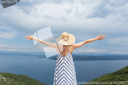 Image of Rear view of young woman wearing striped summer dress and straw hat standing in super bloom of wildflowers, relaxing with hands up to the sky, enjoing beautiful view of Adriatic sea nature, Croatia