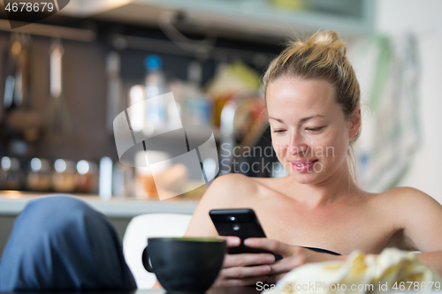 Image of Young smiling cheerful pleased woman indoors at home kitchen using social media on mobile phone for chatting and staying connected with her loved ones. Stay at home, social distancing lifestyle.
