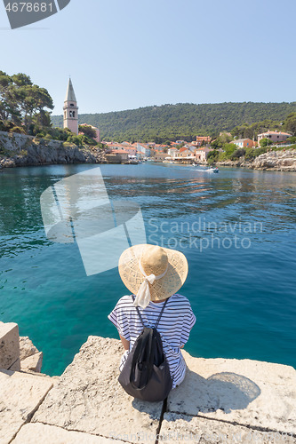 Image of Woman traveler wearing straw summer hat and backpack, sittingat edge of stone pier, enjoying beautiful panoramic view of Veli Losinj, Losinj island, Croatia.