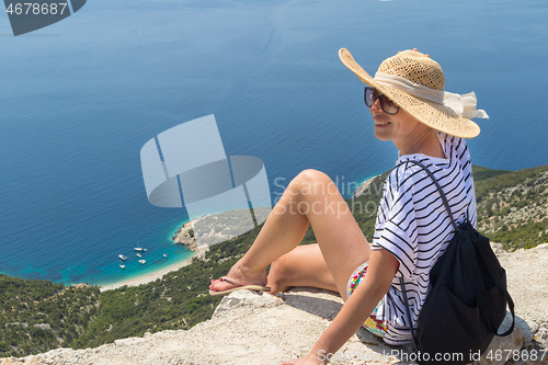 Image of Active sporty woman on summer vacations sitting on old stone wall at Lubenice village, wearing straw hat and beach backpack enjoying beautiful coastal view of Cres island, Croatia