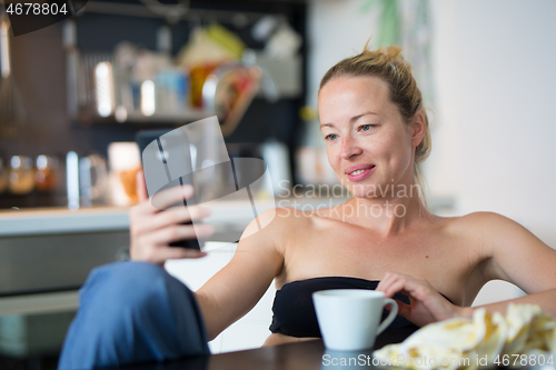 Image of Young smiling cheerful pleased woman indoors at home kitchen using social media on mobile phone for chatting and staying connected with her loved ones. Stay at home, social distancing lifestyle.