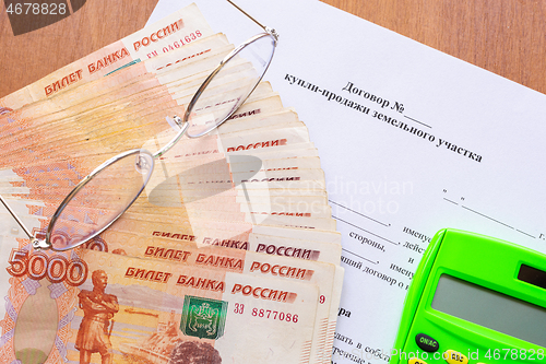 Image of On the table there is a contract for the sale of a land plot, a bundle of money, a calculator and glasses