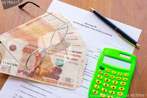 Image of On the table is a contract for the sale of a land plot, a bundle of money, a calculator, a pen and glasses