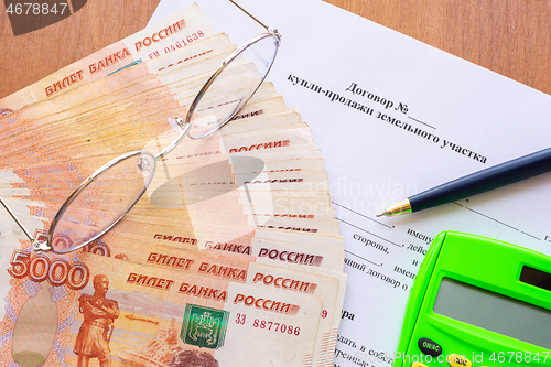 Image of On the table is a contract for the sale of a land plot, a bundle of money, a calculator, glasses and a pen