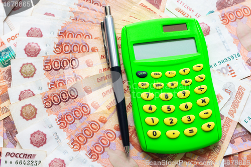 Image of A green calculator and a ballpoint pen lie on a fanned stack of five thousandth bills