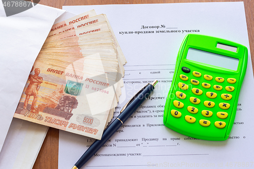 Image of On the table is a contract for the sale of a land plot, a bundle of money in an envelope, a pen and a calculator