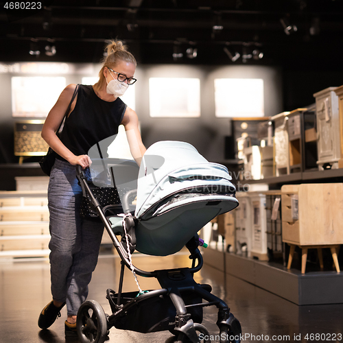 Image of Young mom with newborn in stroller shopping at retail furniture and home accessories store wearing protective medical face mask to prevent spreading of corona virus. New normal during covid epidemic