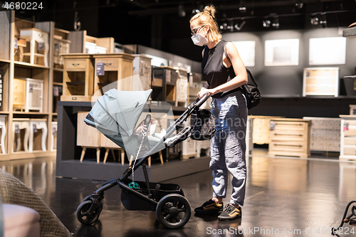 Image of Young mom with newborn in stroller shopping at retail furniture and home accessories store wearing protective medical face mask to prevent spreading of corona virus. New normal during covid epidemic