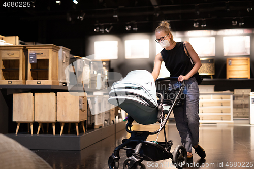 Image of Young mom with newborn in stroller shopping at retail furniture and home accessories store wearing protective medical face mask to prevent spreading of corona virus. New normal during covid epidemic