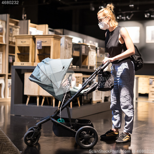 Image of Young mom with newborn in stroller shopping at retail furniture and home accessories store wearing protective medical face mask to prevent spreading of corona virus. New normal during covid epidemic