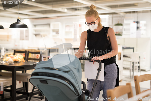 Image of Young mom with newborn in stroller shopping at retail furniture and home accessories store wearing protective medical face mask to prevent spreading of corona virus. New normal during covid epidemic