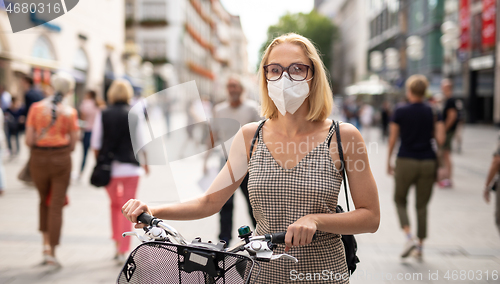 Image of Woman walking by her bicycle on pedestrian city street wearing medical face mask in public to prevent spreading of corona virus. New normal during covid epidemic.
