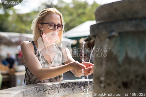 Image of Thirsty young casual cucasian woman wearing medical face mask drinking water from public city fountain on a hot summer day. New life during covid epidemic