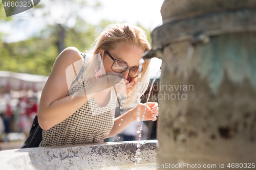 Image of Thirsty young casual cucasian woman wearing medical face mask drinking water from public city fountain on a hot summer day. New social norms during covid epidemic