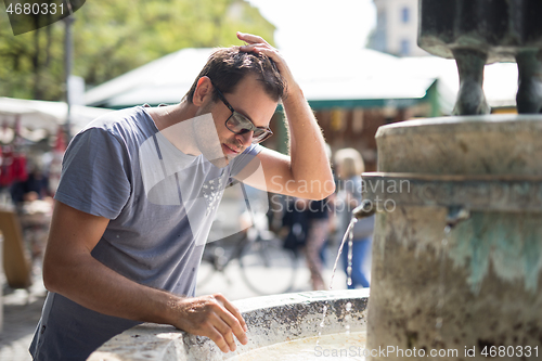 Image of Young casual cucasian man refreshing himself with water from public city fountain on a hot summer day