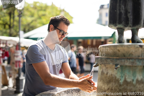 Image of Young casual cucasian man washing hands with water from public city fountain on a hot summer day