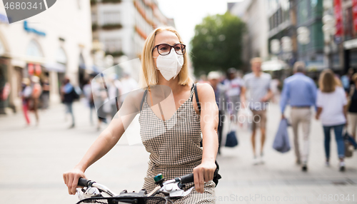 Image of Woman riding bicycle on city street wearing medical face mask in public to prevent spreading of corona virus. New normal during covid epidemic.