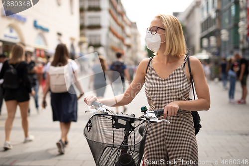 Image of Woman walking by her bicycle on pedestrian city street wearing medical face mask in public to prevent spreading of corona virus. New normal during covid epidemic.