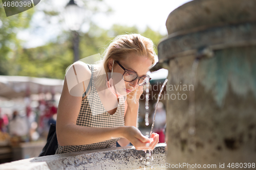 Image of Thirsty young casual cucasian woman wearing medical face mask drinking water from public city fountain on a hot summer day. New social norms during covid epidemic
